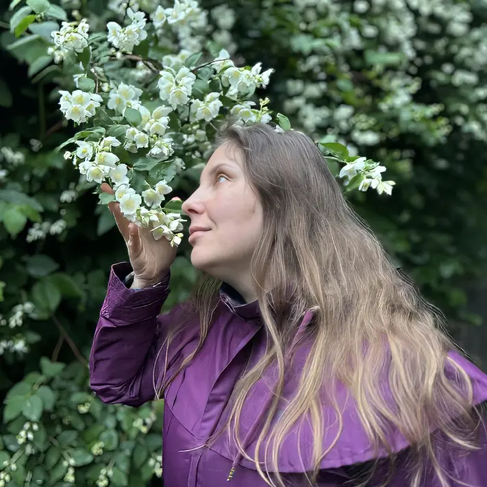 Kwadratowy portret. Magdalena Rutkowska w fioletowej kurtce stoi pośród zieleni i drobnych, białych kwiatów jaśminu. Unosi gałązkę ku twarzy i zwraca ku niej głowę; kwitnący krzew szczelnie wypełnia tło.