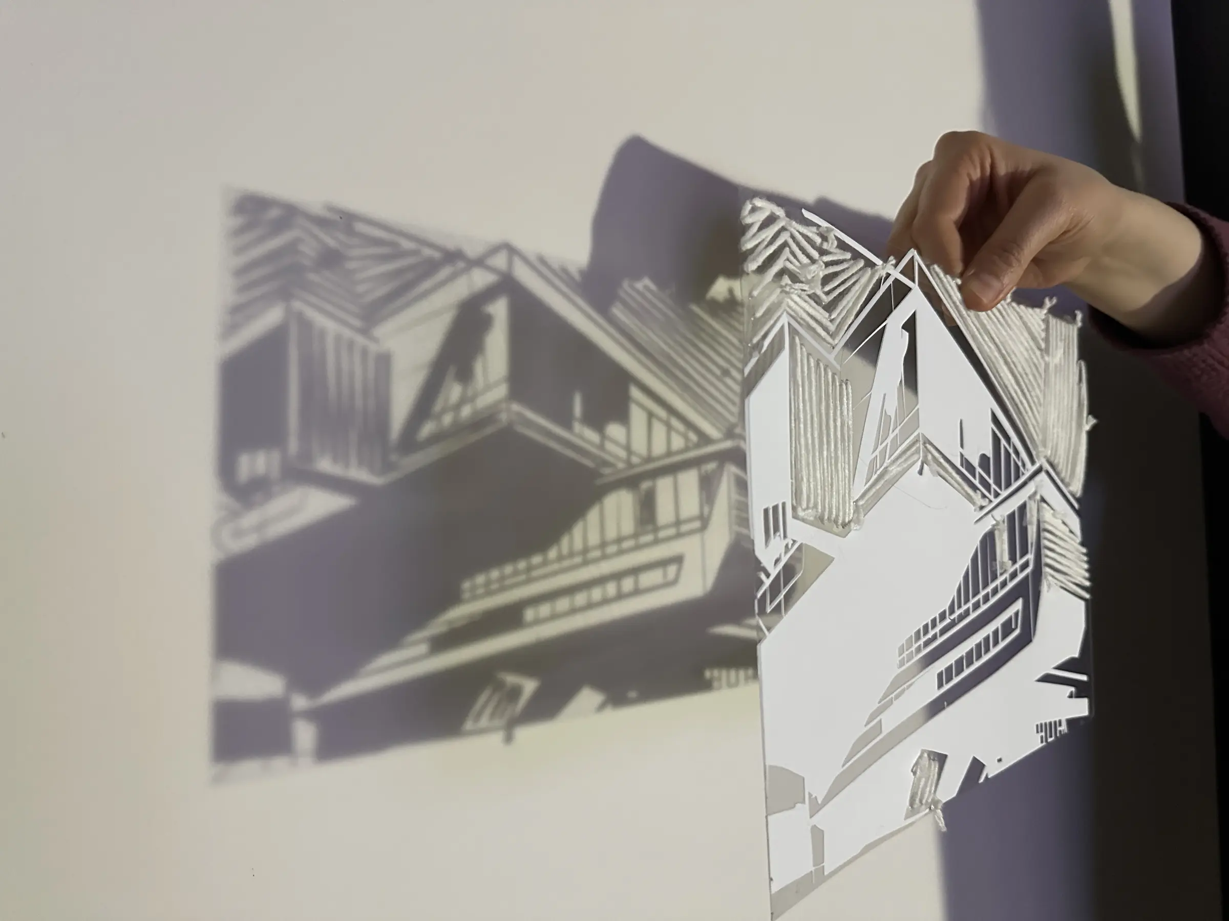 Dłoń trzyma przezroczysty rysunek architektury przed jasną ścianą. Biała materialna formatka zajmuje prawą część kadru, a po lewej pojawia się jej duży, ciemny cień, w którym bryły i pasma przeszyć stają się masywniejsze i lekko rozmyte.