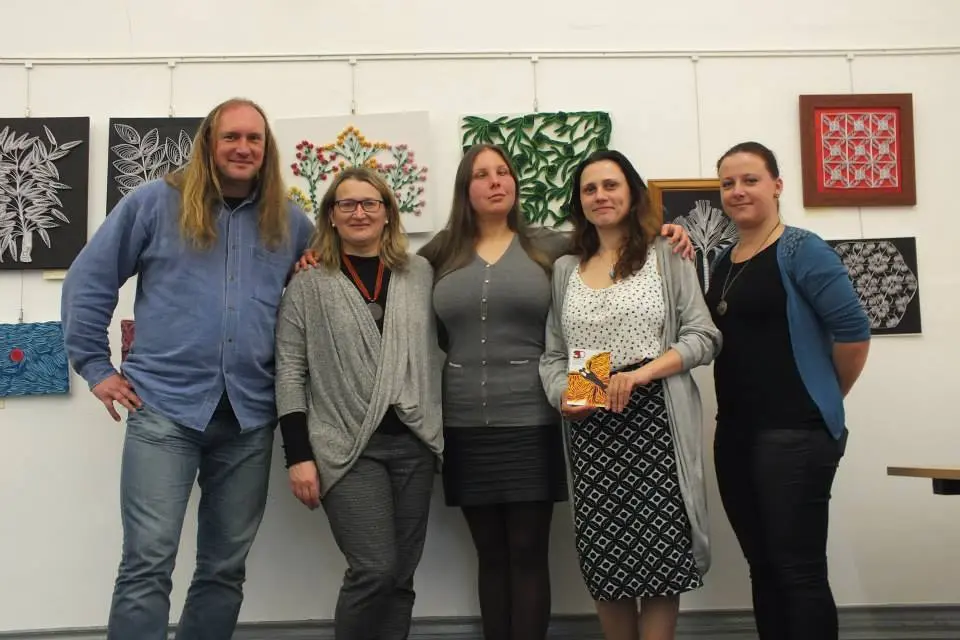 Pięć osób stoi przed ścianą z reliefowymi pracami Magdaleny Rutkowskiej w Falls Road Library. W tle widać czarno-białe kompozycje roślinne oraz mniejsze barwne reliefy.