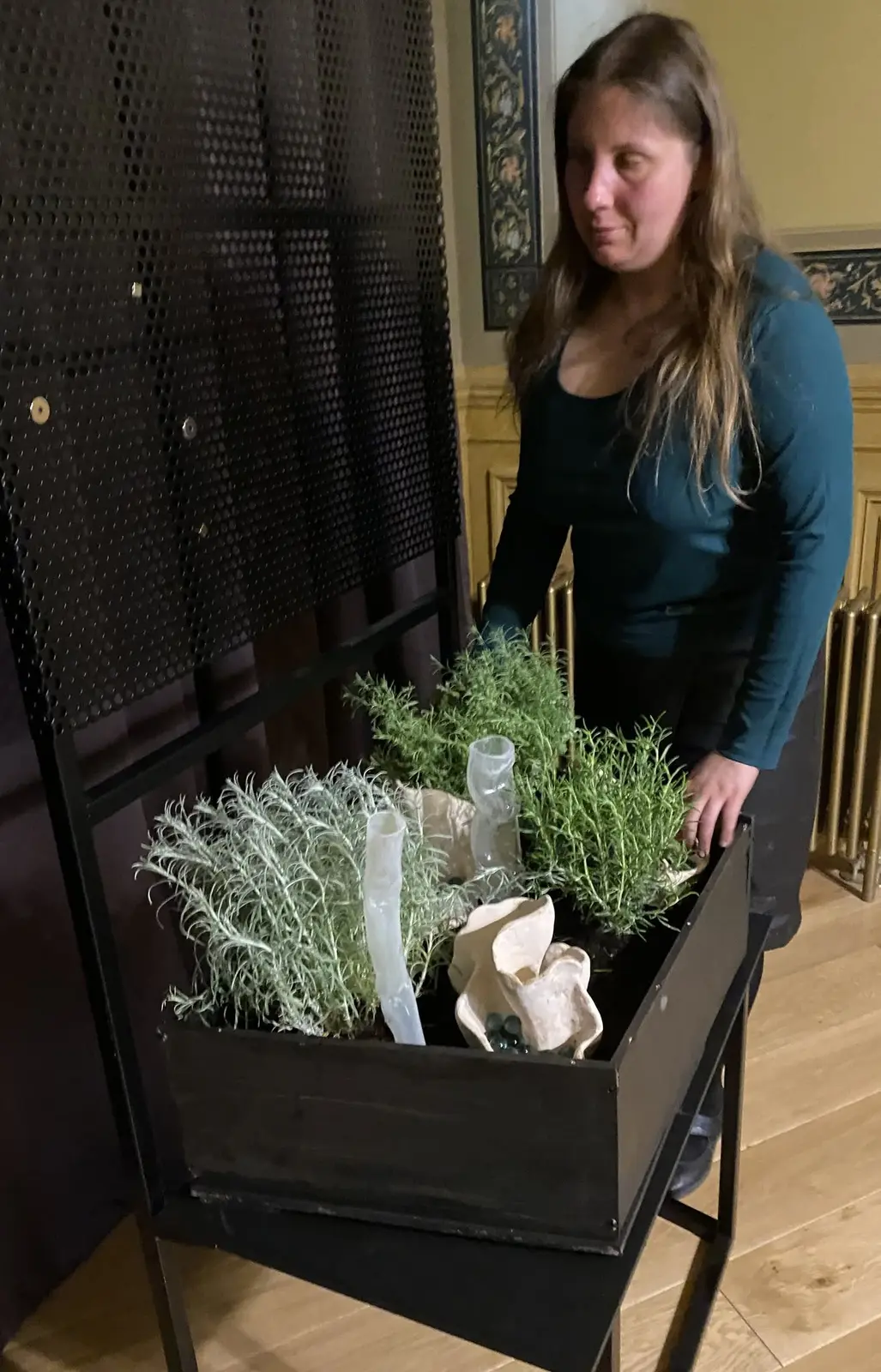 Magdalena Rutkowska stoi obok Ogródka ustawionego w wysokiej, czarnej prostokątnej konstrukcji. Wśród gęstych ziół wyrastają jasne formy ceramiczne i pionowe elementy ze szkła; za instalacją widać ceglaną ścianę i okno Kamienicy Hilarego Majewskiego.