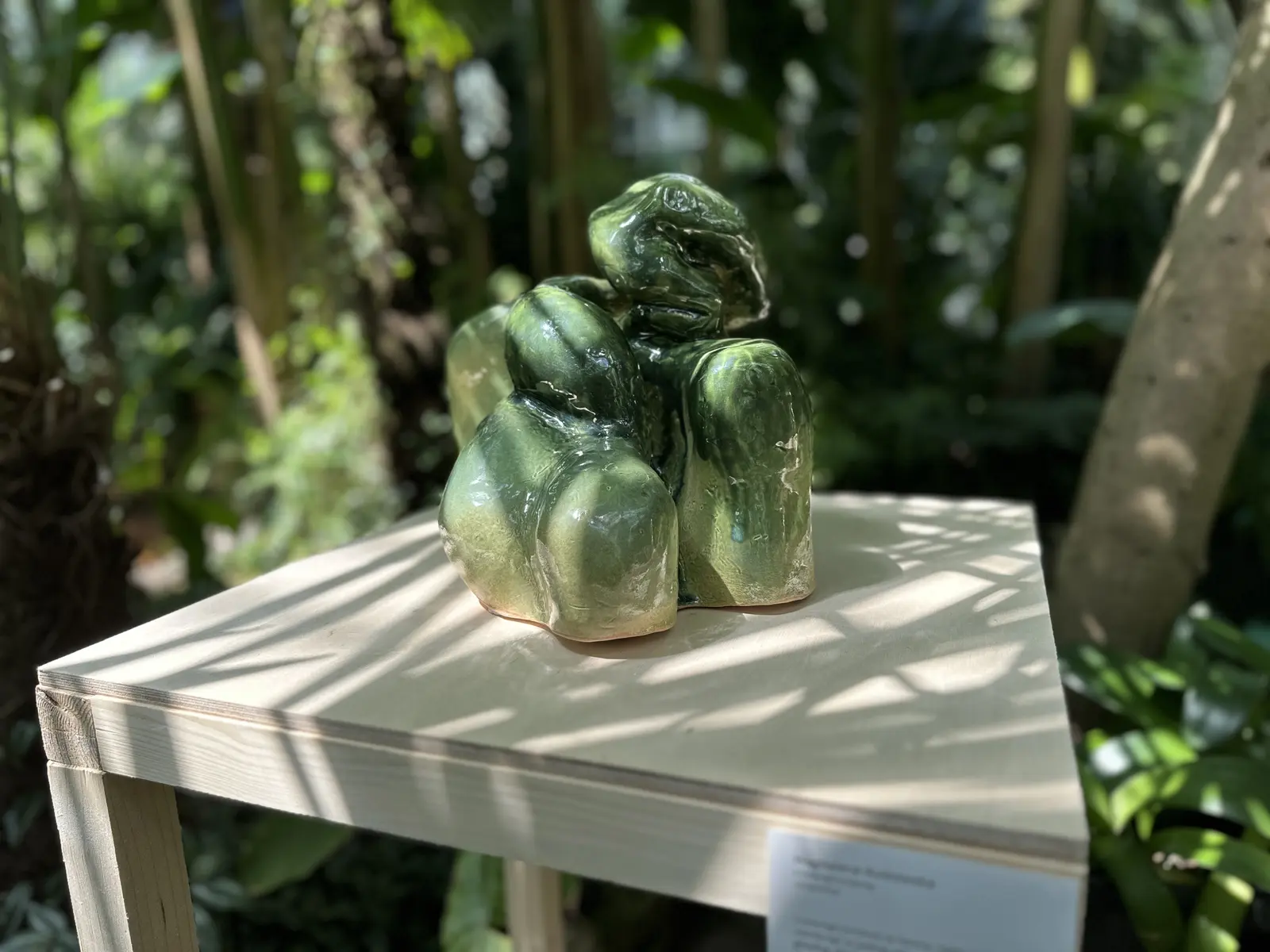 Zbliżenie zielonej, organicznej bryły Biomorfola na jasnym postumencie. Prześwity, refleksy i cienie liści łączą powierzchnię rzeźby z otaczającą ją roślinnością.