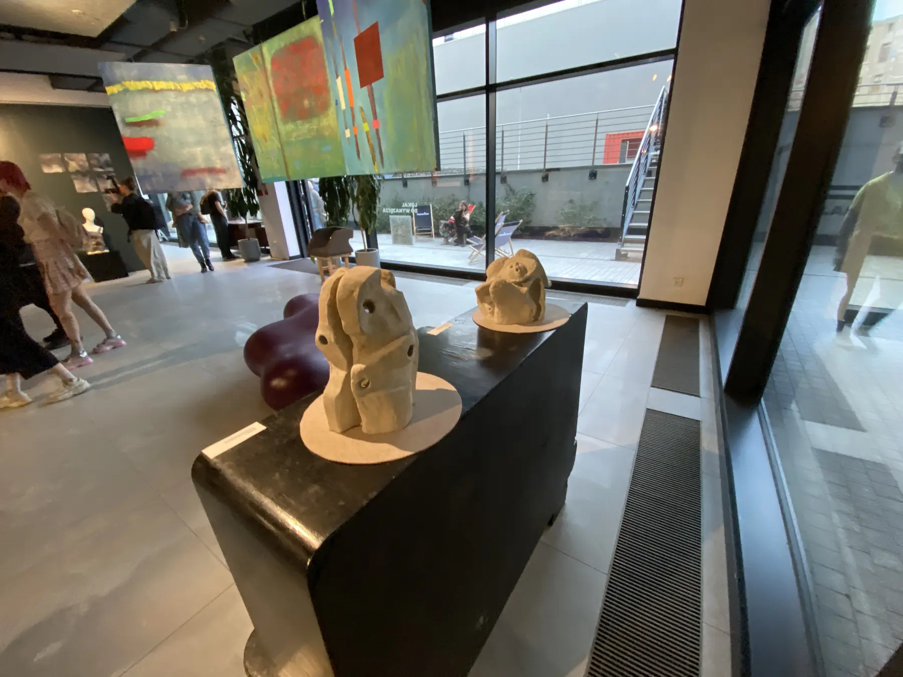 Szeroki widok wnętrza Galerii Pop-up. Na pierwszym planie stoją dwie jasne rzeźby na czarnych postumentach, nad nimi wiszą dwa barwne obrazy, a przez przeszkloną ścianę widać dziedziniec i osoby odwiedzające wystawę.