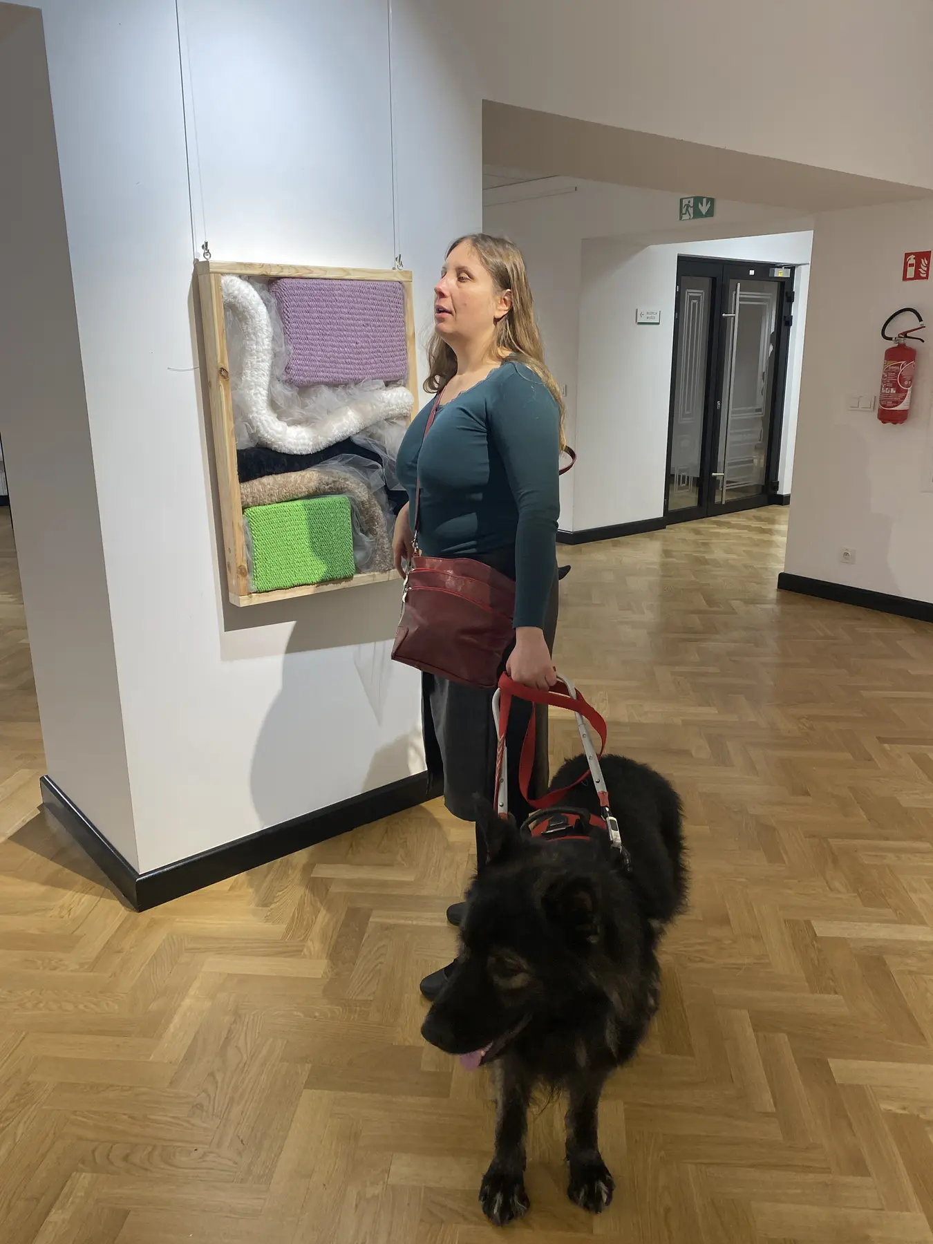 Magdalena Rutkowska stoi z dużym czarnym Pinotem w jasnym korytarzu Akademickiego Centrum Designu. Za nimi wisi Test Lustra: kwadratowa kompozycja z trzema jasnymi, miękkimi formami na polach fioletowym, beżowym i zielonym.