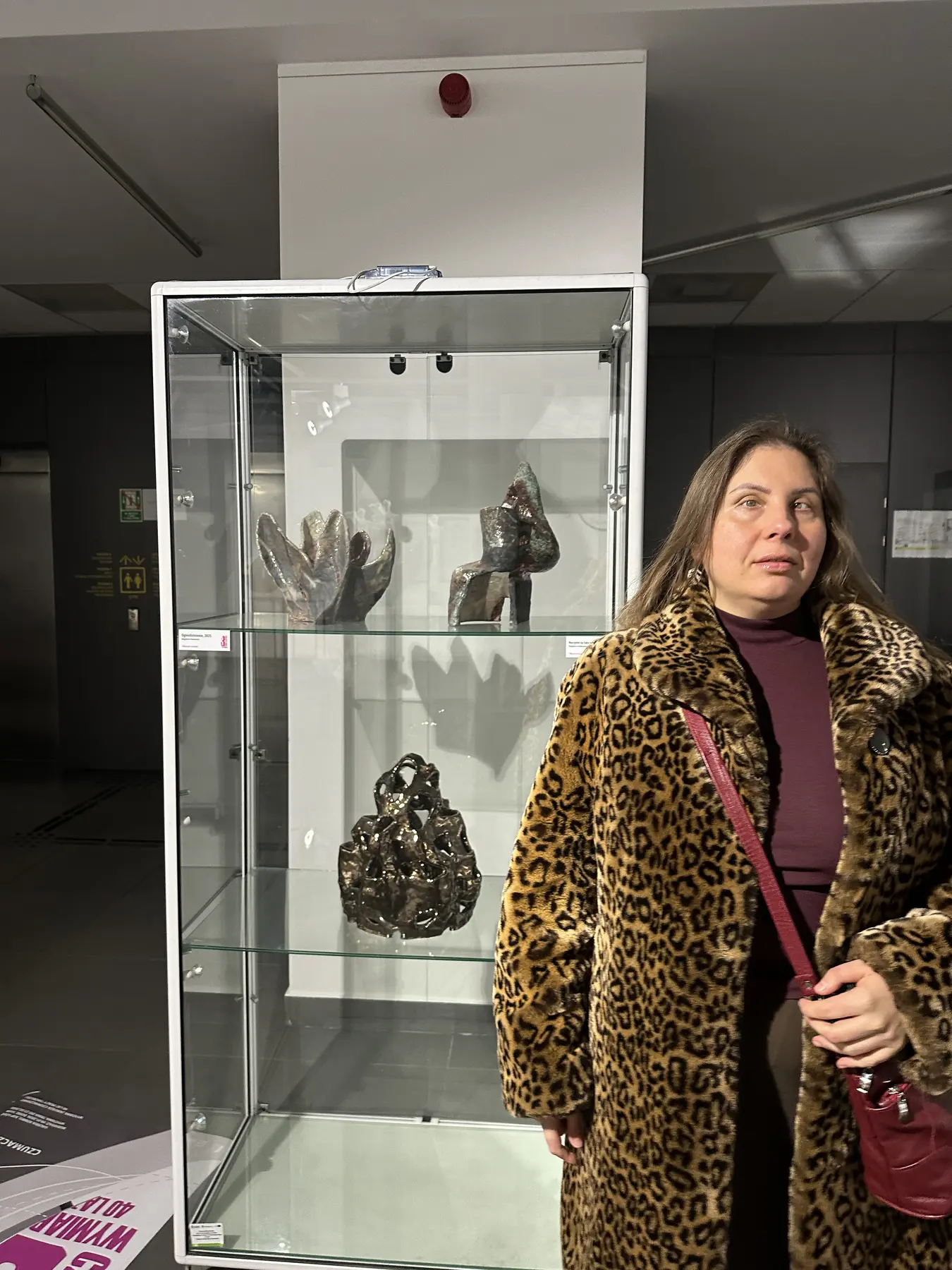 Magdalena Rutkowska stoi przy wysokiej przeszklonej gablocie. Na jej półkach prezentowane są trzy organiczne rzeźby ceramiczne, a wyżej smukła szklana statuetka.