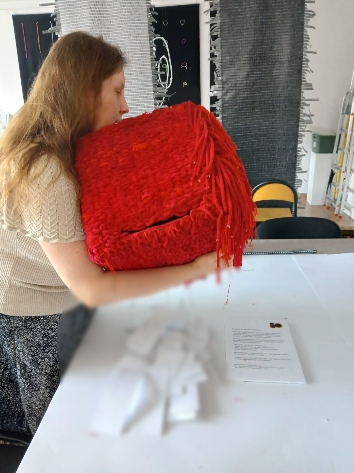 Magdalena trzyma czerwony sześcian przy tułowiu. Ku odbiorcy skierowana jest ściana z poziomą szczeliną, a przy prawym narożniku zwisają frędzle; na stole leżą kartki o nieczytelnej treści.
