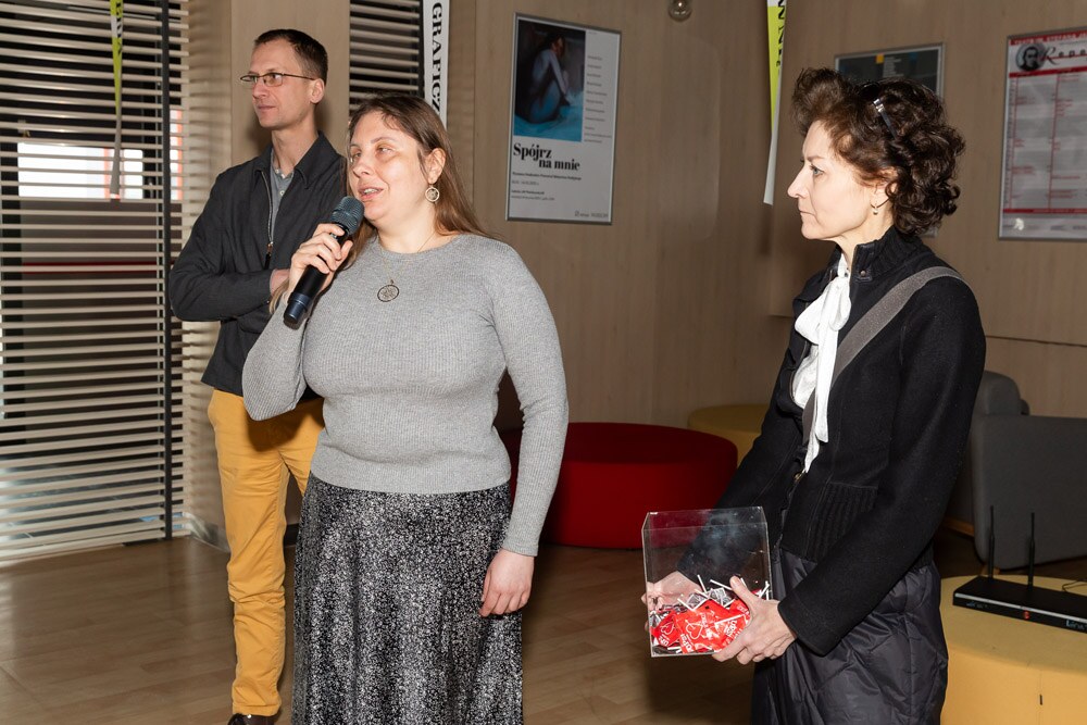 Magdalena mówi do mikrofonu podczas spotkania w holu ASP. Po obu jej stronach stoją dwie osoby; jedna trzyma przezroczysty pojemnik z czerwonymi przedmiotami.