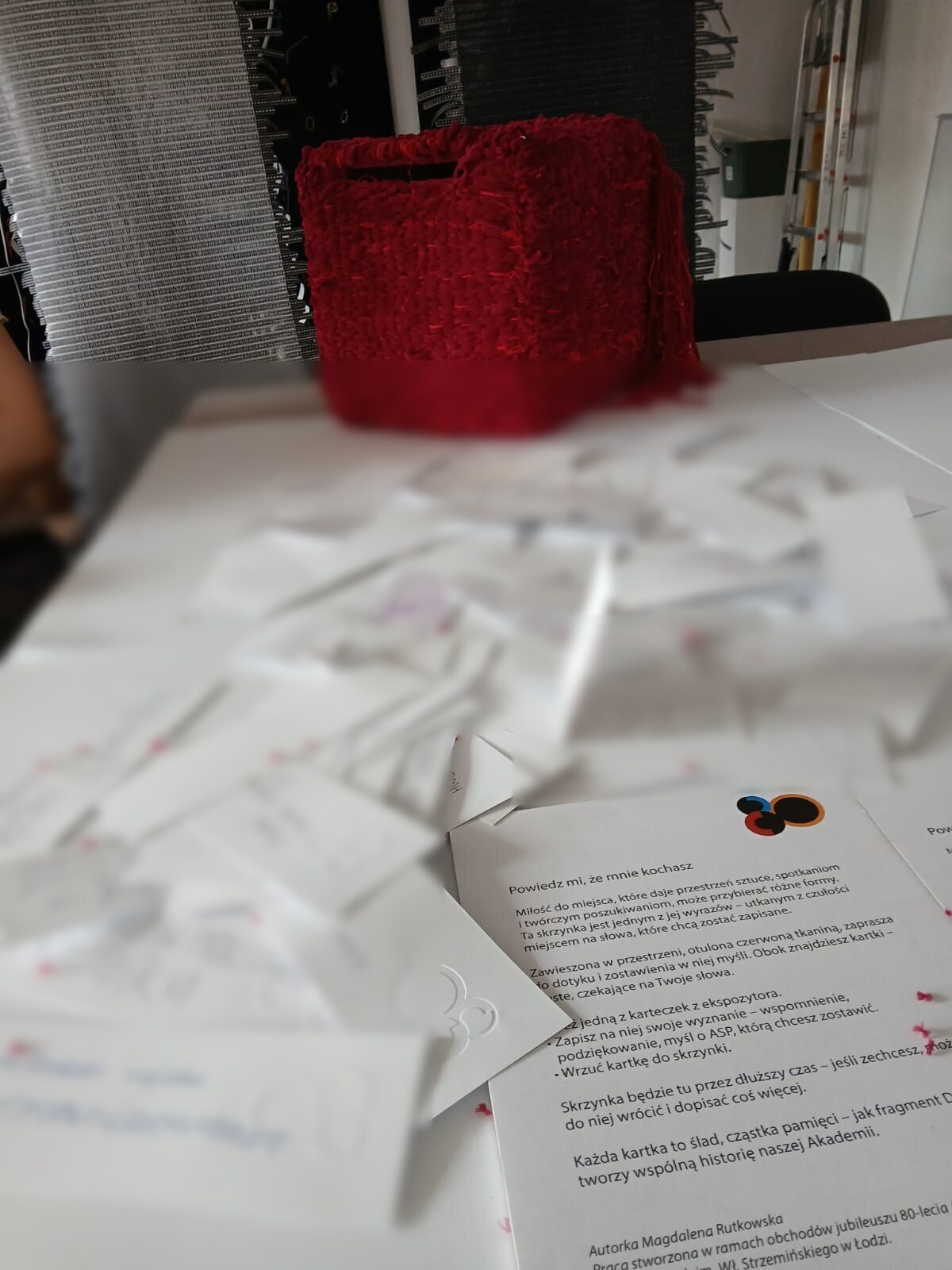 Czerwony sześcian stoi na końcu stołu pokrytego dziesiątkami białych kartek wydobytych z jego wnętrza. Na pierwszym planie leży drukowana instrukcja; prywatne zapisy na kartkach są nieczytelne.