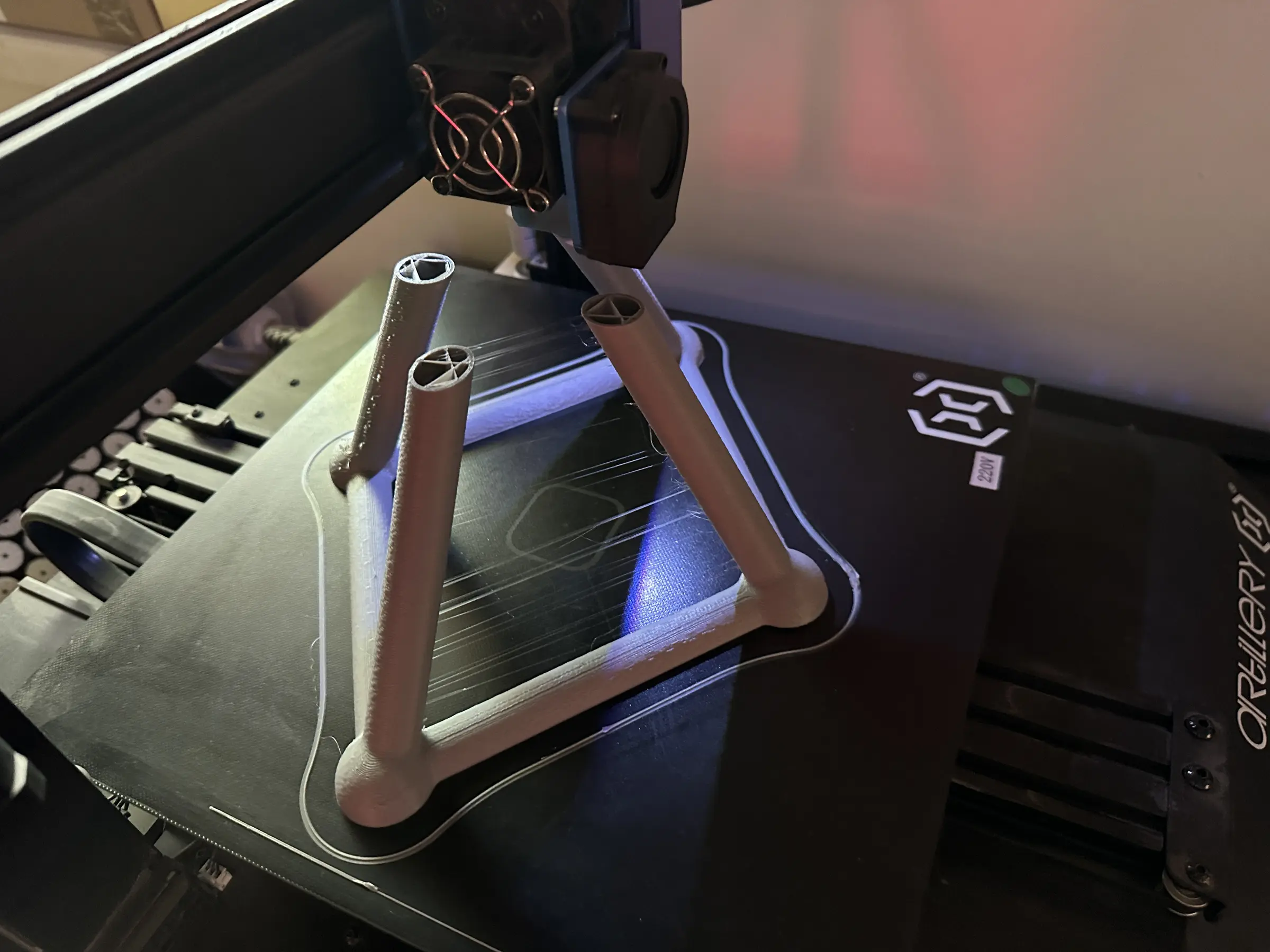 Zbliżenie wnętrza drukarki 3D. Na czarnym stole roboczym powstaje szara przestrzenna konstrukcja z zaokrąglonymi węzłami i trzema ukośnie wznoszącymi się prętami. Ponad niedokończonym szkieletem znajduje się czarna głowica drukująca.