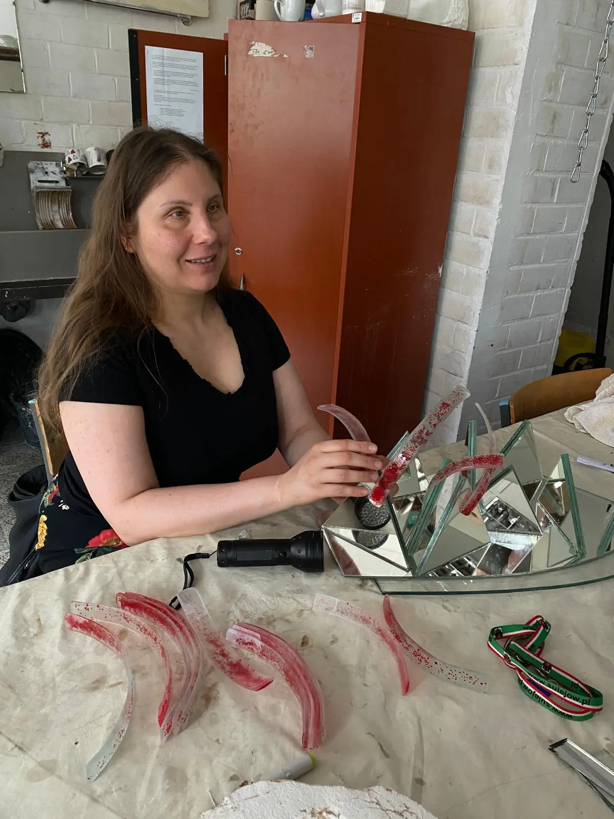 Magdalena Rutkowska siedzi przy pracownianym stole, lekko uśmiechnięta, i trzyma oburącz wygięty pas przejrzystego szkła z czerwonym nakrapianiem nad układem lustrzanych trójkątów. Przed nią leży kilka kolejnych pasm o różnych krzywiznach. Błyszczące powierzchnie rzeźby odbijają szkło, dłonie i otoczenie.