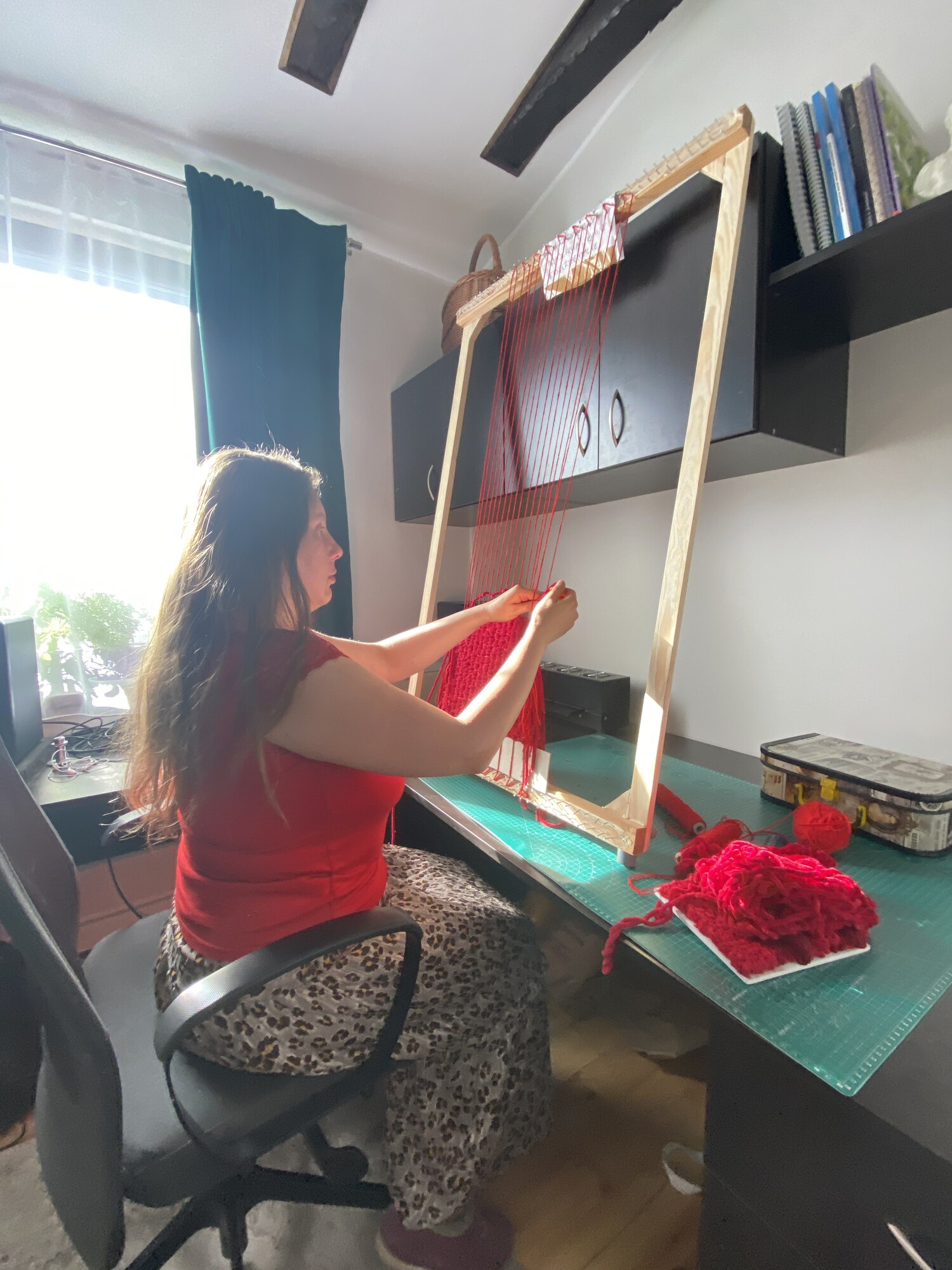 Magdalena siedzi przy wysokiej drewnianej ramie tkackiej w domowej pracowni. Jej dłonie pracują przy czerwonej tkaninie, a na stole leżą kłębki i kolejne fragmenty materiału.