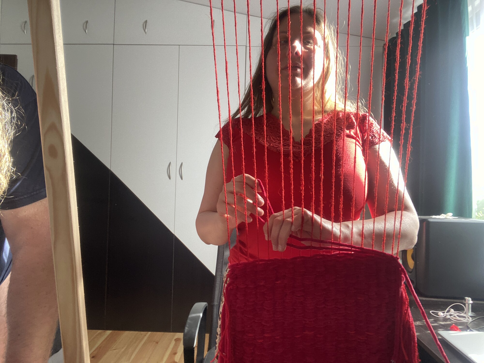 Magdalena widziana przez pionowe czerwone nici osnowy. Jej twarz i dłonie przecinają rytmiczne linie nici, a u dołu kadru znajduje się powstający panel.
