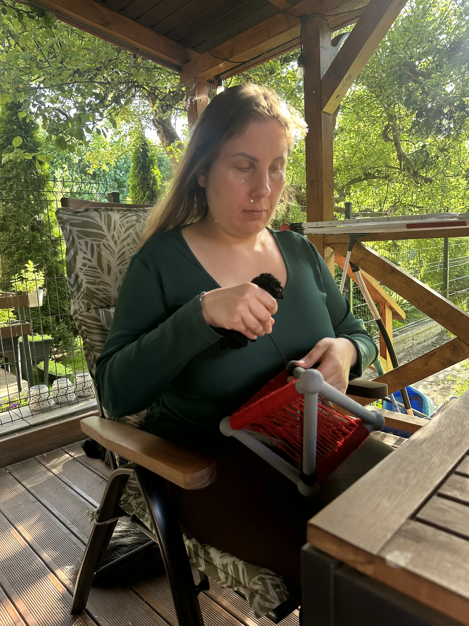 Magdalena siedzi w drewnianym fotelu na zadaszonym tarasie i spogląda na oparty o kolana szary szkielet, częściowo wypełniony intensywnie czerwoną przędzą. Jedną dłonią unosi zwiniętą czarną przędzę, drugą prowadzi ją przy obiekcie.