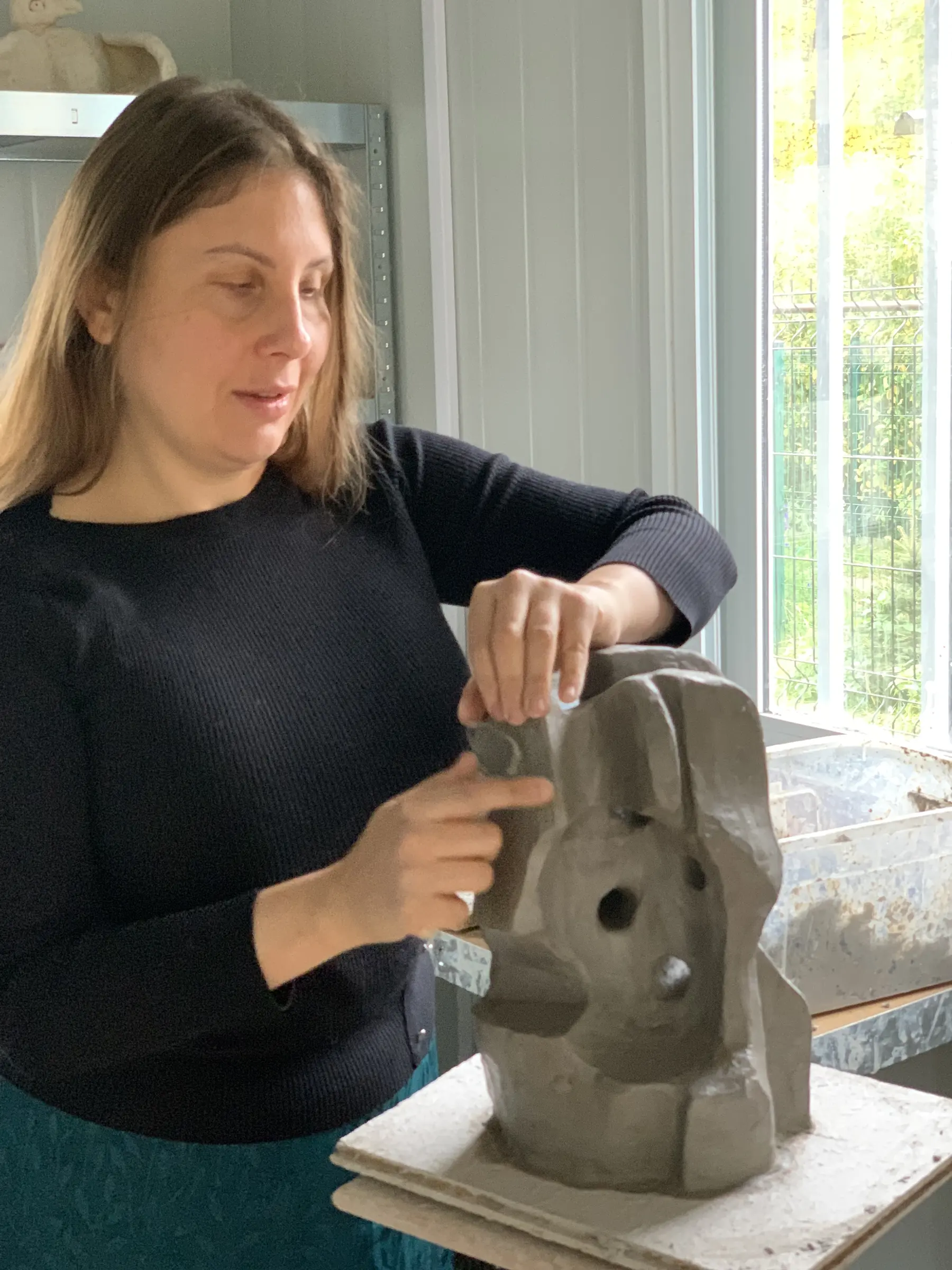 Magdalena pochyla się nad wilgotną, szarą gliną. Jedną dłonią modeluje zaokrągloną górną krawędź, a drugą opracowuje niższą część bryły. Z bliska widać miękkie fałdy materiału, świeże ślady palców i głębokie otwarcie pomiędzy pionowymi masami.