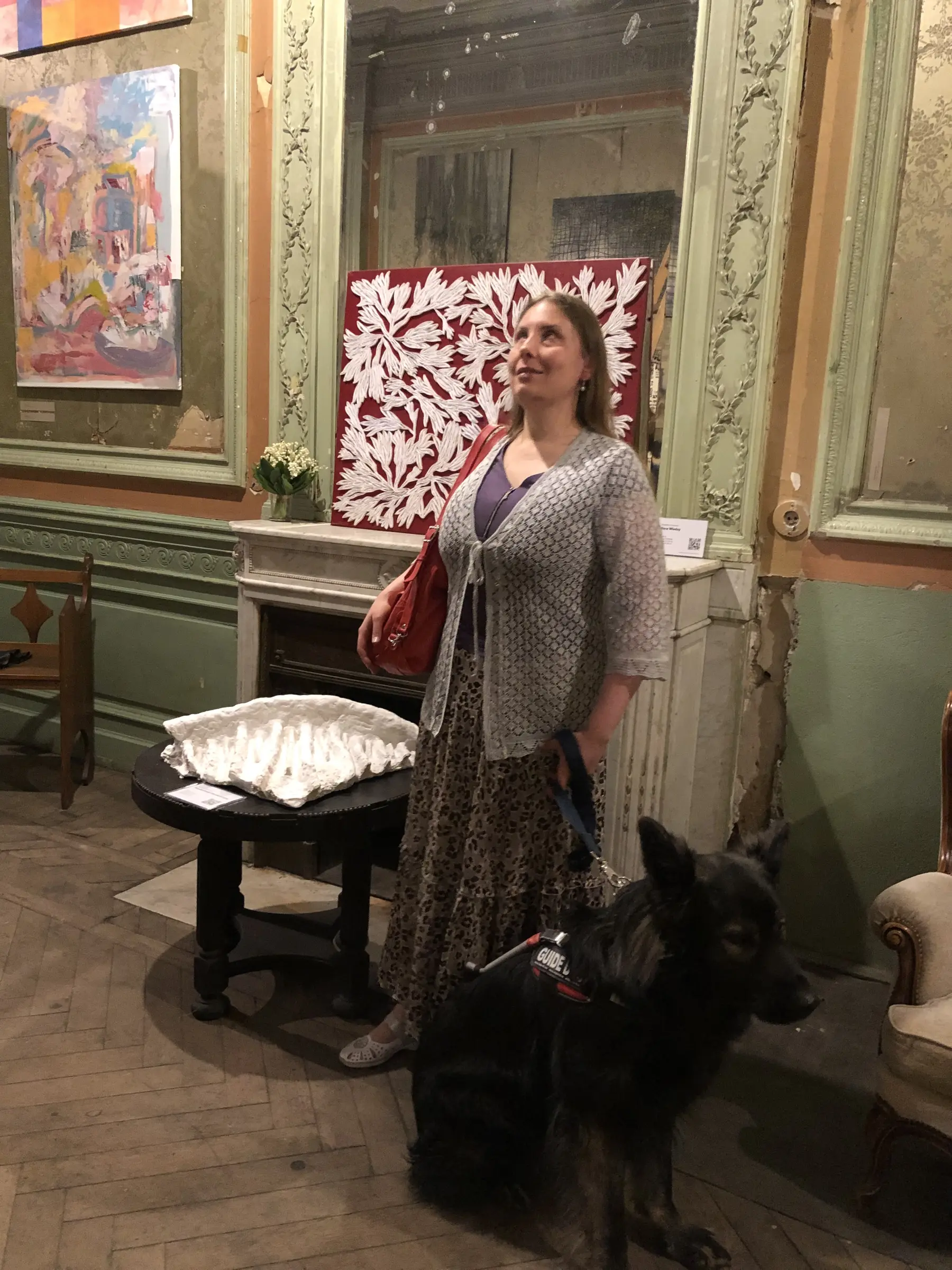 Magdalena stoi przy „Rzeźbotworze Pandemicznym” leżącym na czarnym stoliku. Jest lekko uśmiechnięta, a twarz ma zwróconą ku górze; obok niej, na pierwszym planie, stoi czarny Pinot w uprzęży. W tle widać kominek, ornamentalne zielone ramy i inne dzieła.