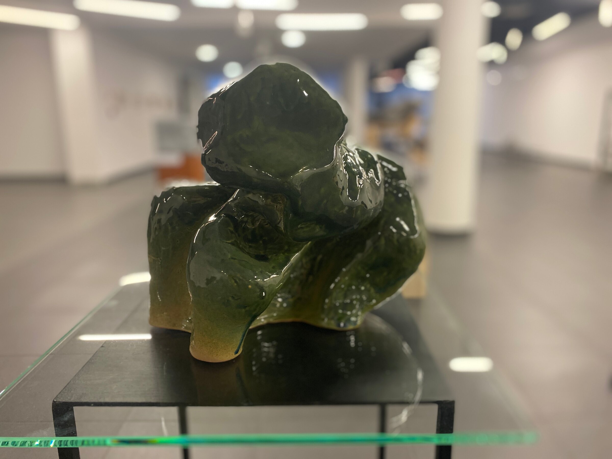 Poziome, bliskie ujęcie zielonej, szkliwionej rzeźby ceramicznej w jasnej galerii. Zwarta organiczna bryła składa się z dużych zaokrąglonych wypukłości, głębokich zagłębień i grubych, skręconych fałd. Czarna płyta i szklany blat są elementami ekspozycji.