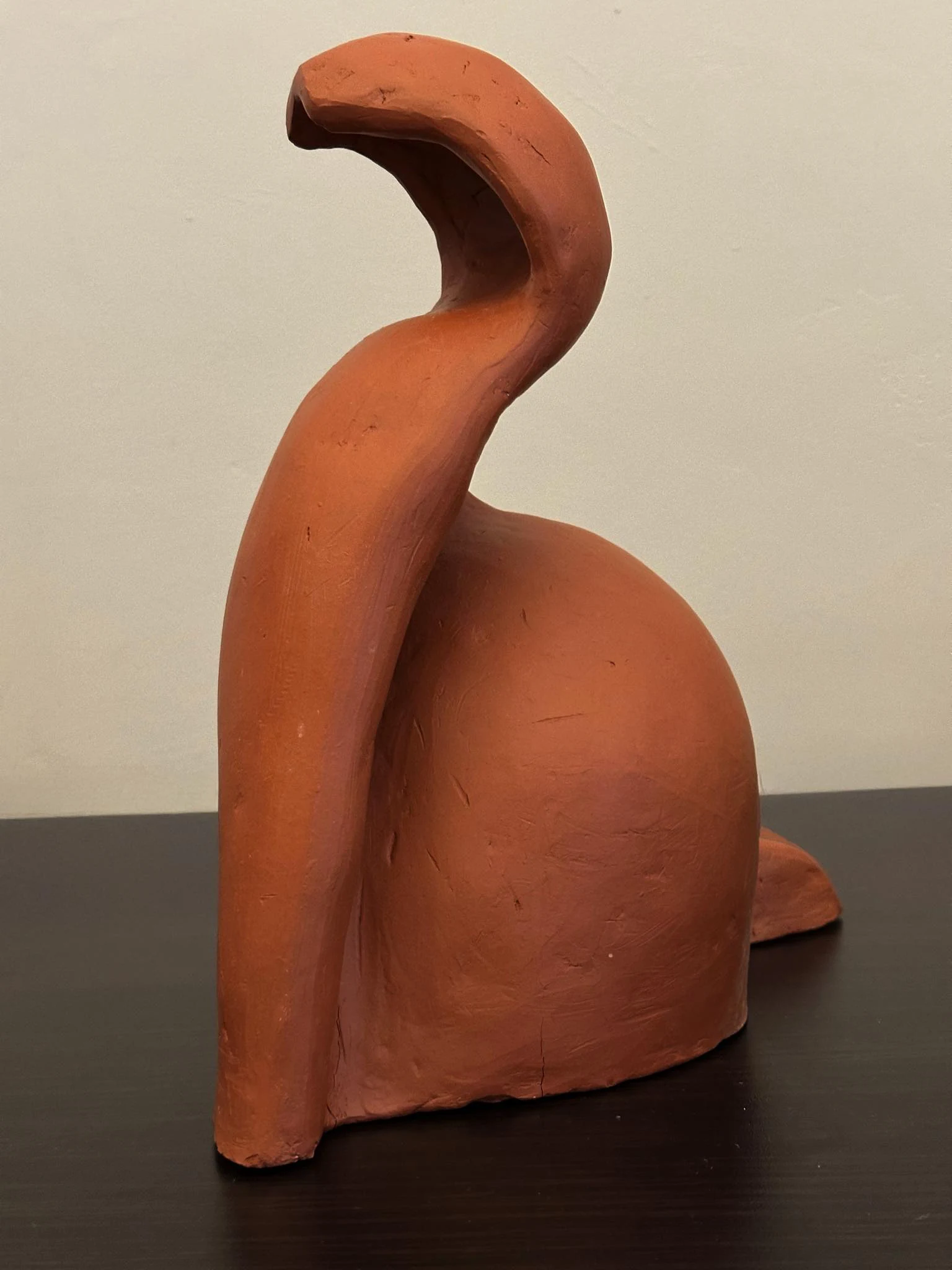 Ceramiczna rzeźba „Nie Kot, Nie Wąż” widziana ukośnie. Z szerokiej, nierównej podstawy wyrasta gruba, lekko spłaszczona partia, która biegnie niemal pionowo, a u góry zawija się nad głębokim zagłębieniem. Po prawej łączy się z dużą, łagodnie wypukłą masą; pomiędzy obiema częściami pozostaje wąska, zacieniona szczelina. Matowa powierzchnia ma na fotografii ciepły, ceglasto-rdzawy kolor oraz widoczne drobne wgłębienia, rysy i ślady modelowania.