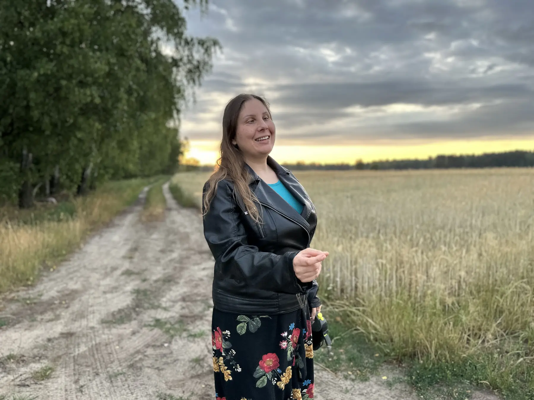 Magdalena Rutkowska stoi na polnej drodze przy łanie zboża i uśmiecha się. Ma na sobie czarną kurtkę skóropodobną i długą czarną spódnicę w kwiaty; za nią rozciąga się wieczorny pejzaż pod zachmurzonym niebem.