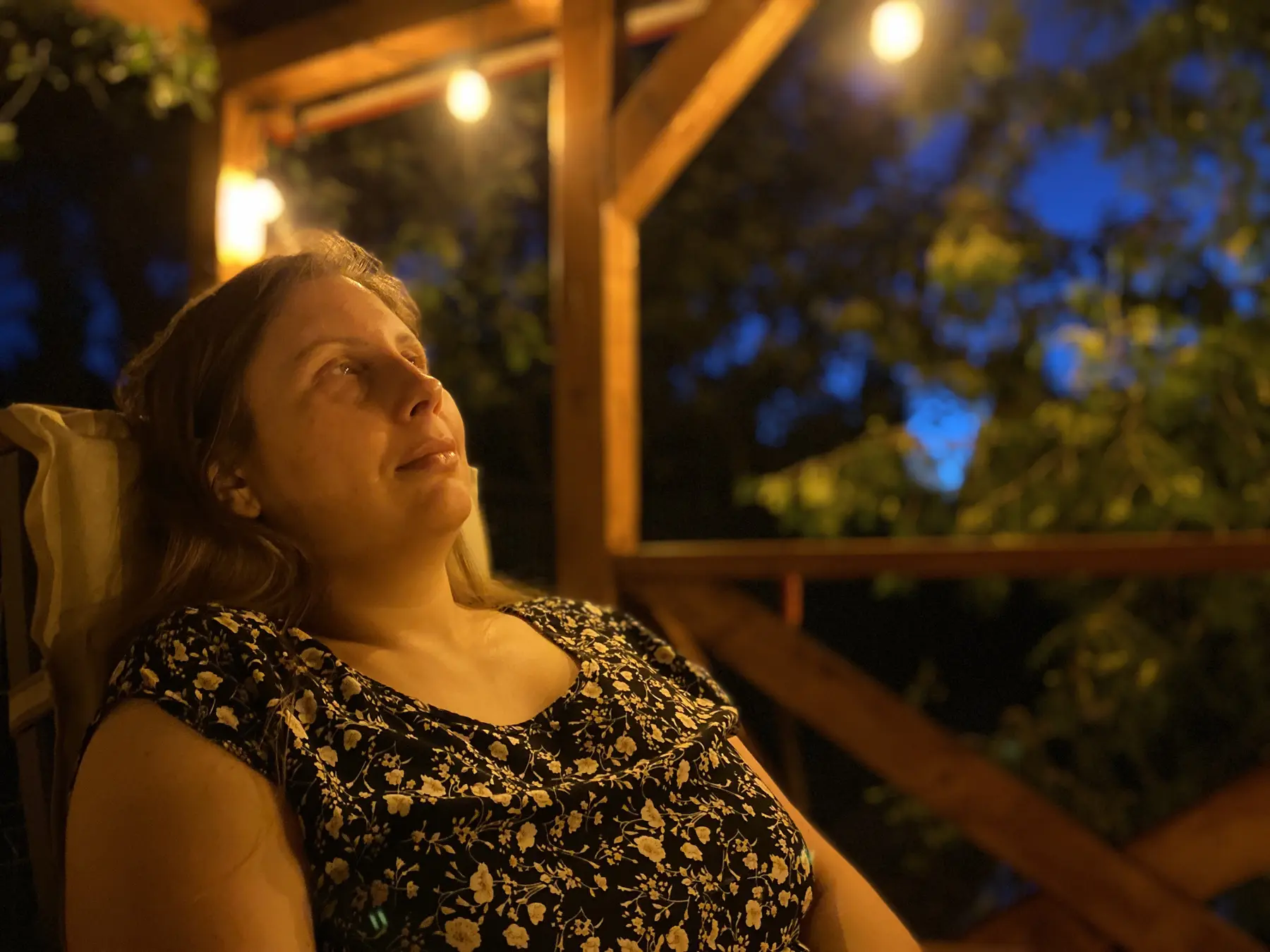 Magdalena Rutkowska odpoczywa wieczorem pod drewnianym zadaszeniem. Ciepłe światło lamp wydobywa jej twarz z ciemności; w tle rozmyte drzewa i głęboki błękit nieba.
