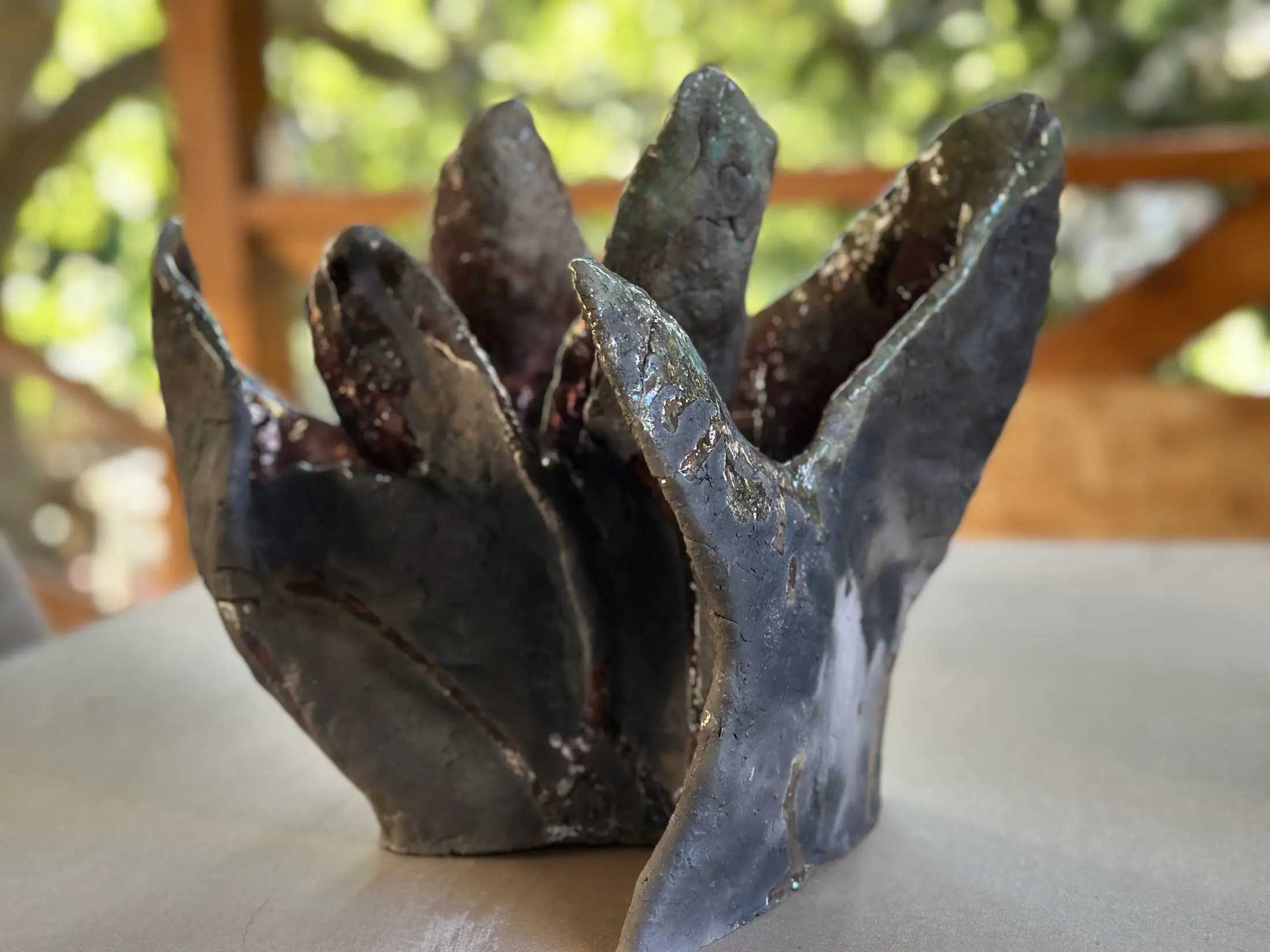 Ceramiczna rzeźba oglądana z przodu i lekko z boku. Wysokie, nieregularne płaty wznoszą się wokół otwartego wnętrza. Ich grafitowo-czarne powierzchnie mają spękania oraz metaliczne refleksy turkusu, miedzi i srebra; między nimi widać ciemne czerwienie i purpury.