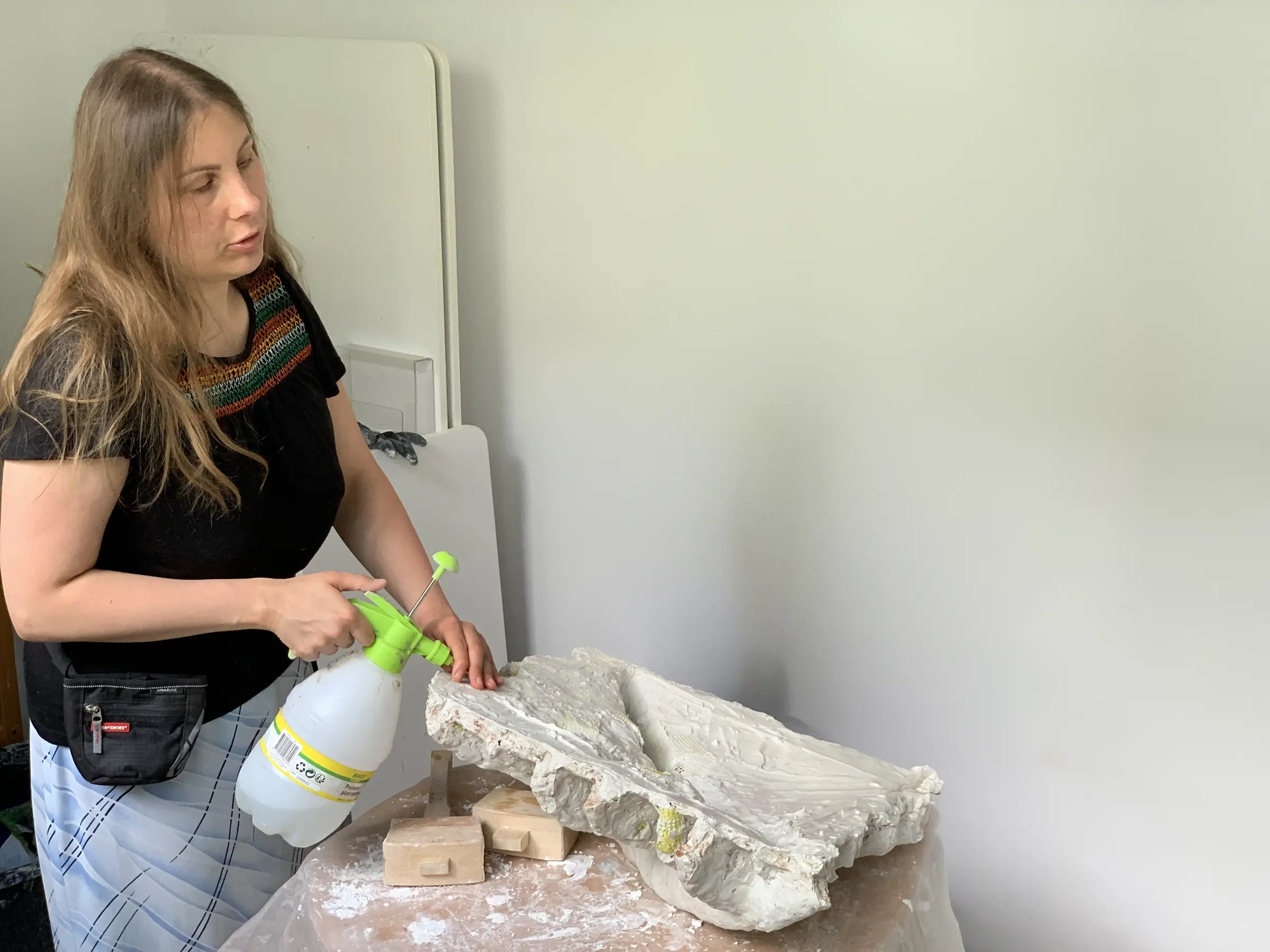 Magdalena stoi przy rozłożonym na stole surowym gipsowym odlewie. Trzyma duży zielony opryskiwacz, a drugą dłonią dotyka nierównej krawędzi rzeźby; w powierzchni widać brązowawe ślady i fragmenty żółtej siatki.