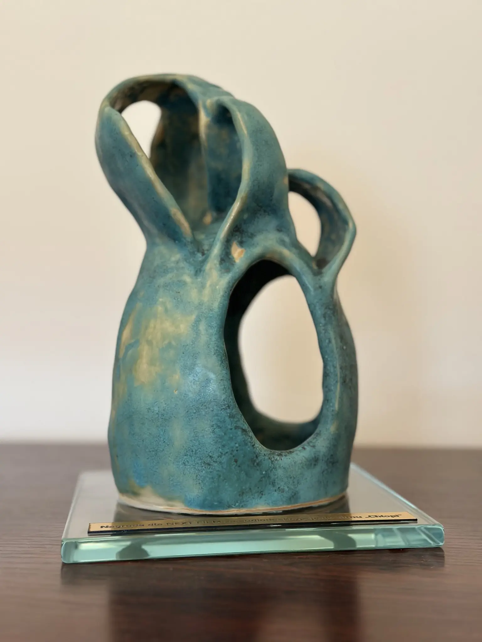 Turkusowo-zielona statuetka ceramiczna na kwadratowej szklanej podstawie, oglądana lekko z góry i z boku. Zwarta bryła rozszerza się ku dołowi, a wyżej przechodzi w kilka szerokich, łukowo wygiętych pasm. Pomiędzy nimi otwierają się nieregularne prześwity: największy, pionowy, zajmuje dolną prawą część formy, mniejsze pojawiają się wyżej i po bokach. Szkliwo jest niejednolite, z kremowo-ochrowymi przejaśnieniami i ciemnym nakrapianiem. Do podstawy przymocowana jest metalowa tabliczka.