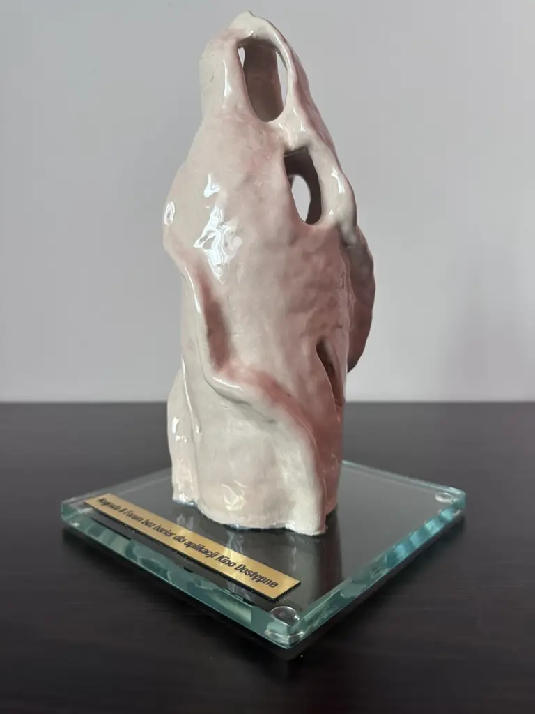 Ukośny widok statuetki ukazujący jej nieregularną głębokość. Szerokie partie ceramiczne nakładają się, wybrzuszają i zwężają, tworząc duży otwór w górnej części, owalny prześwit pośrodku oraz wąską szczelinę niżej. Błyszczące szkliwo przechodzi od mlecznej bieli do bladego różu i ciemniejszych różowoszarych smug. Szklana podstawa oraz przymocowana tabliczka są widoczne w perspektywie.