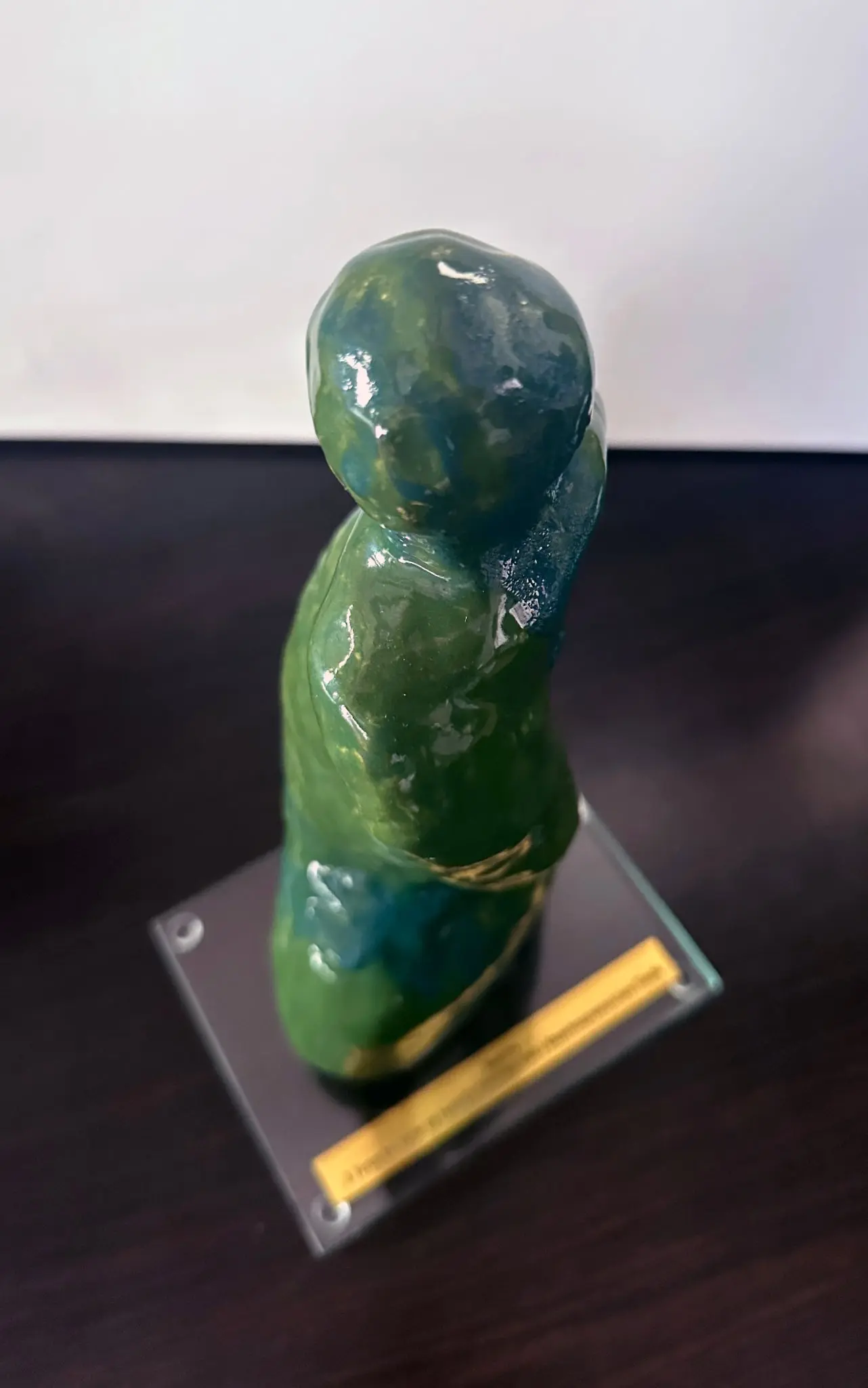 Ta sama statuetka oglądana z góry i z ukosa. Zaokrąglona głowa jest przesunięta względem zwężonej szyi, a jedna z górnych partii biegnie ukośnie wzdłuż boku, ujawniając skręt i nieregularną głębokość bryły. Zieleń szkliwa jest jaśniejsza w świetle, z ciemnym niebiesko-granatowym pasmem po prawej stronie i licznymi białymi refleksami na nierównej powierzchni. Pod formą widać ukośny obrys prostokątnej szklanej podstawy oraz rozmytą metalową tabliczkę.
