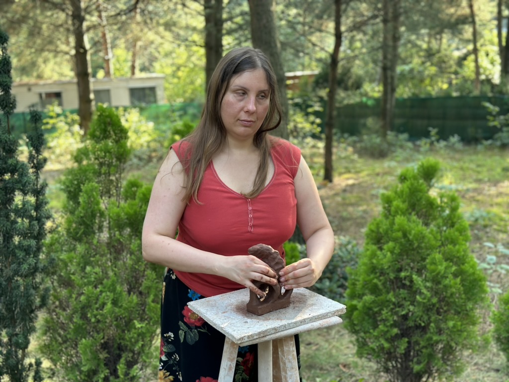 Magdalena Rutkowska modeluje glinianą rzeźbę na kawalecie ustawionym wśród drzew. Ma na sobie czerwony top i ciemną spódnicę w kwiaty.