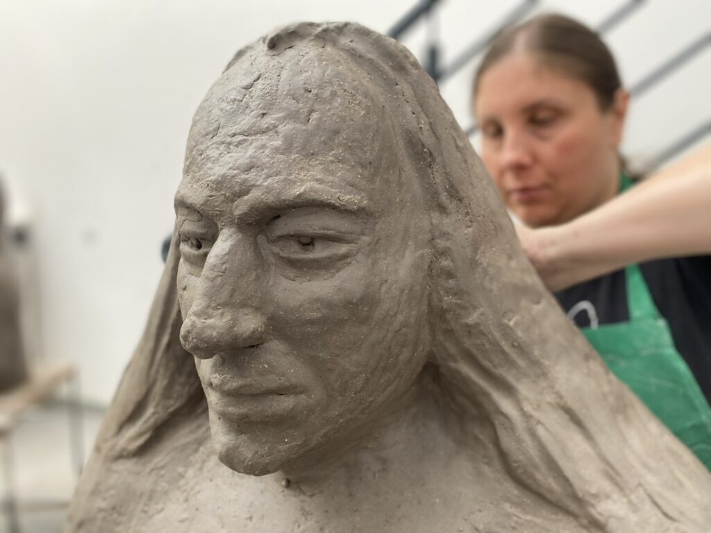 Magdalena Rutkowska pracuje nad dużą glinianą rzeźbą głowy mężczyzny z długimi włosami. Jej postać widoczna jest w tle, za rzeźbą.