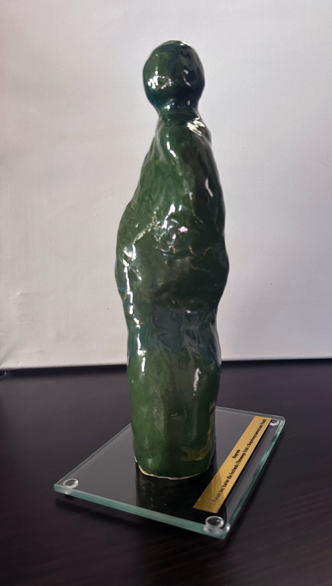 Widok boczny smukłej statuetki o organicznej, antropomorficznej sylwecie. Głowa jest niewielka i lekko pochylona, tułów wydłużony i subtelnie skręcony, a dolna część bardziej masywna. Połyskująca powierzchnia łączy odcienie zieleni i granatu, które przenikają się na całej formie. Rzeźba ustawiona jest na prostokątnej szklanej podstawie ze złotą tabliczką.
