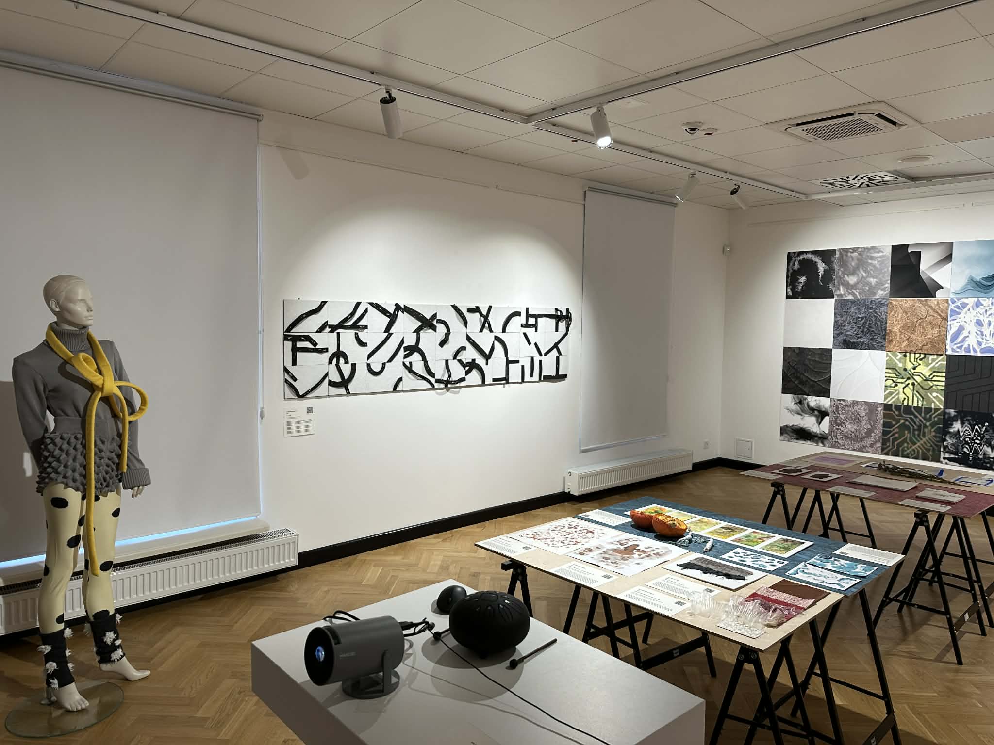 Widok większego fragmentu sali wystawowej. Na środkowej ścianie wisi długa, pozioma kompozycja: czarne, dynamicznie wygięte pasy biegną nad wydłużonym białym tłem. W przestrzeni stoją stoły z materiałami edukacyjnymi i obiektami do oglądania. Po lewej stronie widoczny jest manekin w szaro-żółtej stylizacji, a po prawej na sąsiedniej ścianie duża kompozycja złożona z wielu kwadratowych paneli o różnych fakturach i kolorach.