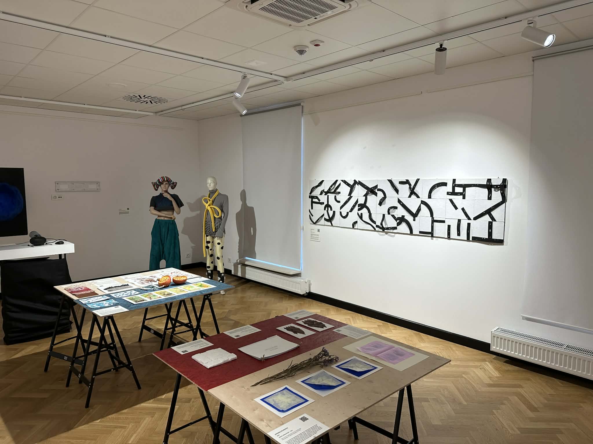 Szeroki widok fragmentu sali wystawowej. Na białej ścianie po prawej wisi długa, pozioma kompozycja ścienna: na wydłużonym białym tle biegną czarne, wygięte pasy o dynamicznych kształtach. Przed ścianą stoją dwa stoły ekspozycyjne z materiałami, próbkami i planszami. Po lewej stronie widać manekin ubrany w eksperymentalny strój z żółtym, pętlącym się elementem na torsie. Sala ma jasne ściany, parkiet w jodełkę i sufit z reflektorami.