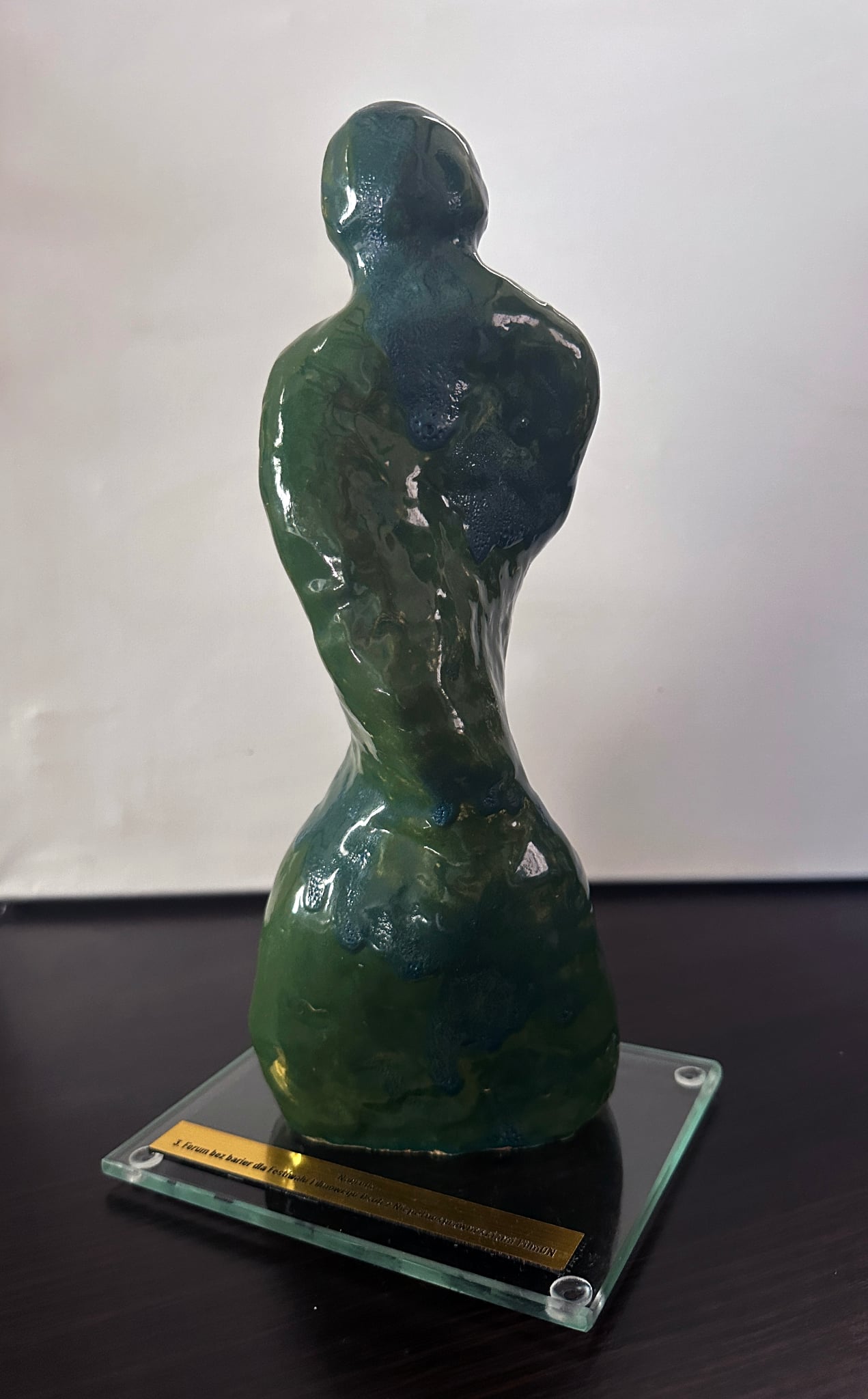 Połyskująca statuetka o uproszczonej ludzkiej formie, ukazana z przodu i lekko z boku, z delikatnie skręconą osią ciała. Głowa i tułów nie są ustawione frontalnie, co nadaje sylwetce wrażenie ruchu. Rzeźba jest smukła, w odcieniach zieleni i ciemnego błękitu z miękkim, marmurkowym przenikaniem barw, ustawiona na prostokątnej szklanej podstawie ze złotą tabliczką.