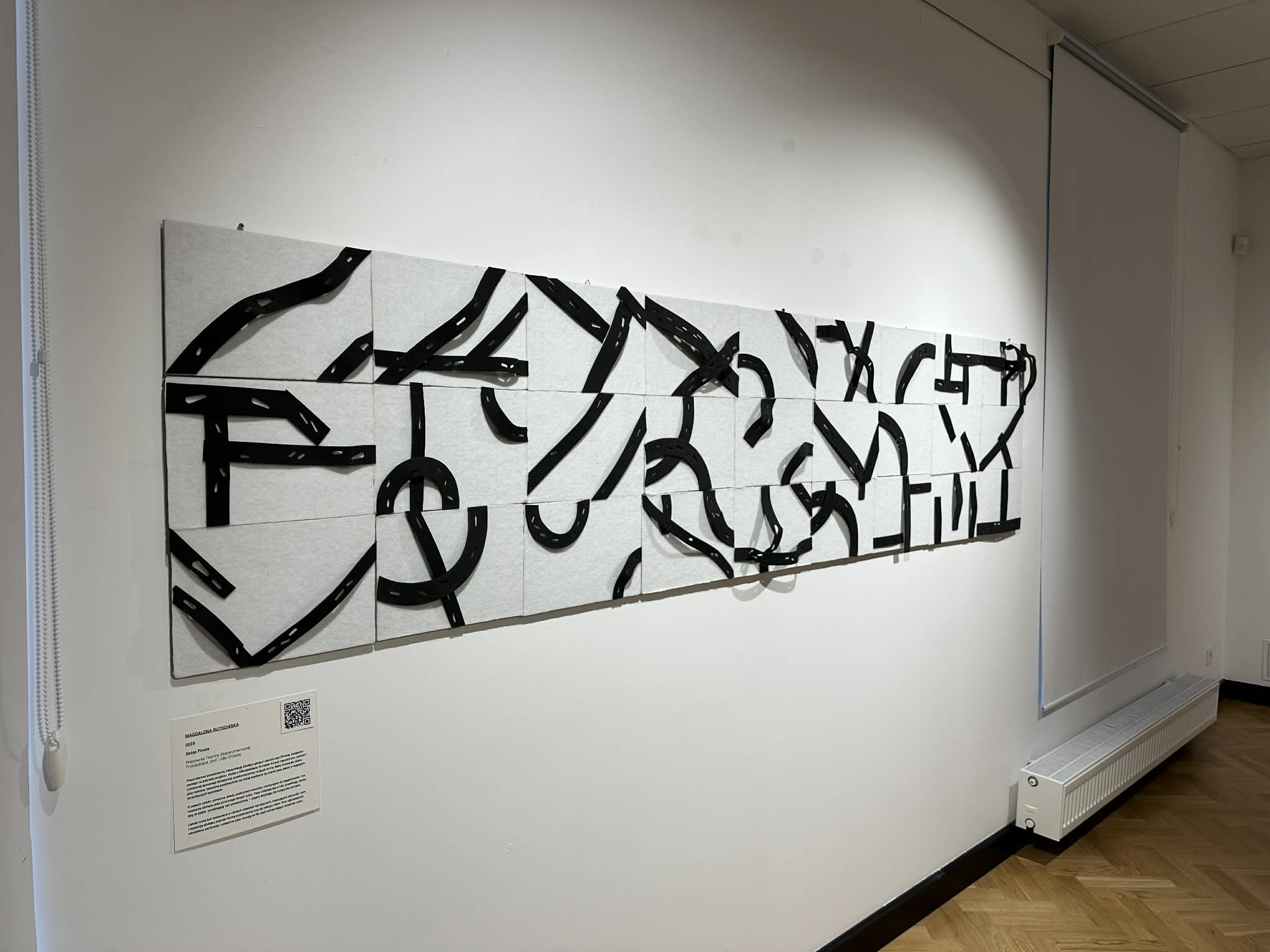 Zbliżenie pracy „Galop Pinota” Magdaleny Rutkowskiej. To długa, pozioma kompozycja ścienna na białym tle. Tło składa się z prostokątnych, filcowych formatek ułożonych w dwóch rzędach. Nad ich powierzchnią przebiegają czarne pasy pianki o wygiętych i łamanych kształtach, z rytmicznymi podłużnymi otworami. Pasy układają się w łuki, przenikają się i miejscami dotykają tła tylko końcami. Pod pracą znajduje się tabliczka z opisem i kodem QR.