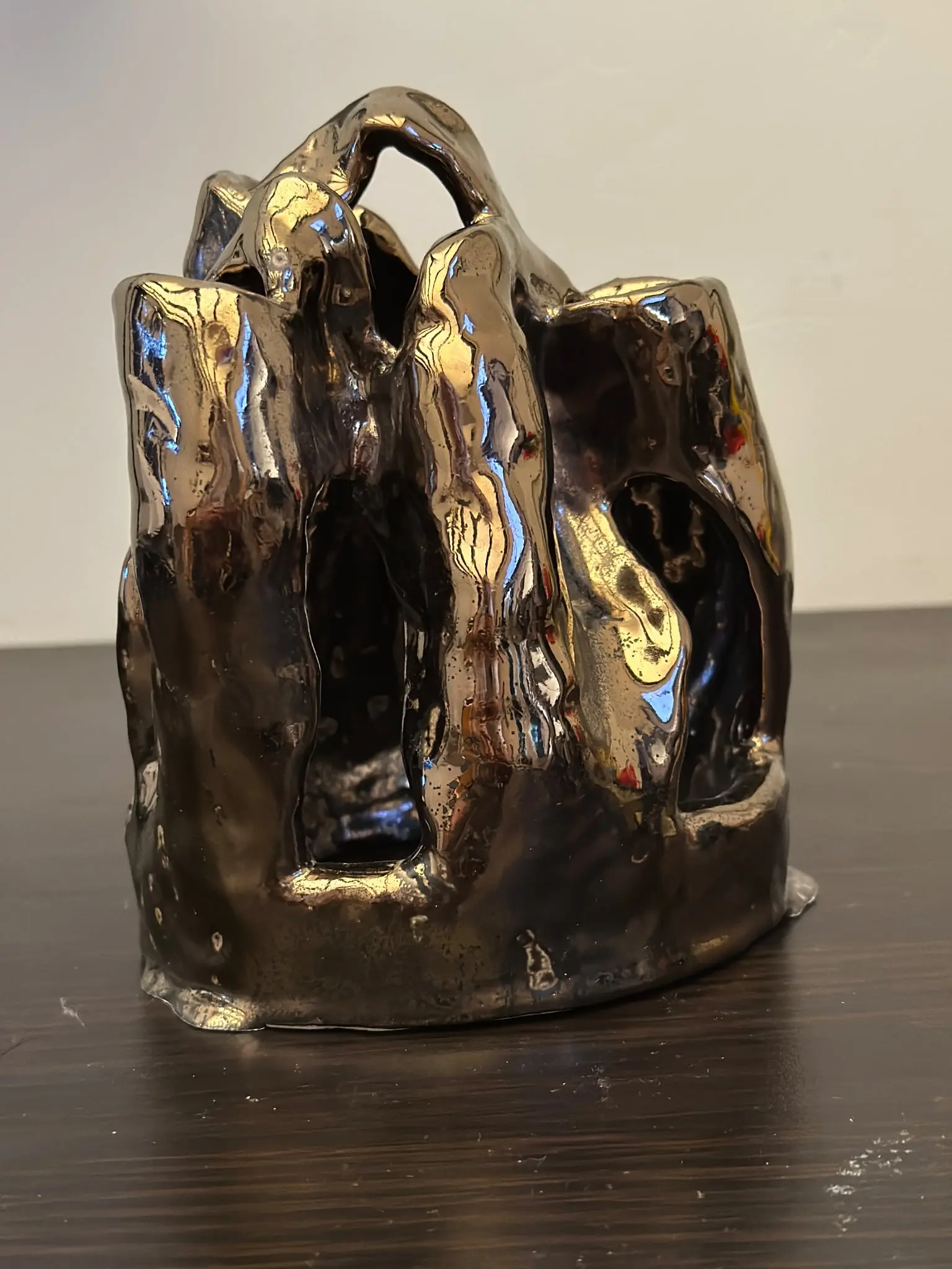 Mniejsza rzeźba ceramiczna pokazana osobno, stojąca na ciemnym drewnianym blacie przed jasną ścianą. Z niskiej, nieregularnej podstawy wznosi się kilka grubych ścian i płatów różnej wysokości, połączonych u góry łukowymi mostami. Pomiędzy nimi znajdują się duże, pionowe otwory. Błyszczące metaliczne szkliwo przechodzi od ciemnego brązu i czerni do złotych pól oraz wąskich niebieskich refleksów.